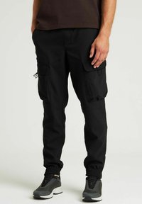 Pantalons cargo noirs avec une coupe décontractée, pr�ésentant des poches latérales, des poignets élastiques et une texture de tissu lisse. Portés avec des chaussures de sport grises.