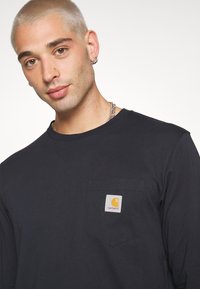 Fekete, hosszú ujjú pamut póló. Elülső zsebe a Carhartt narancs és sárga logójával díszített. Klasszikus környakú fazon.