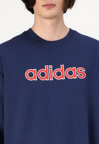 Ung person som bär en marinblå sweatshirt med röd och vit "adidas"-logga centrerad på bröstet.