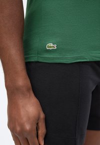 Chemise verte avec petit logo de crocodile brodé près de la taille, portée avec un short noir, montrant une main gauche à la peau foncée reposant le long du corps.