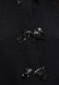 Svart tröja med en texturerad stickad yta, med tre tygrosetter som har paljetter i varierande positioner på framsidan.