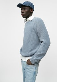 Gai zila ribota džemperis, uzlikts uz balta krekla ar apkakli, kopā ar gaiša denima džinsiem. Izskatu pabeidz teksturēta zilā cepure.