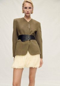 Olive green blazer with structured shoulders, belted waist, and black belt. Paired with a cream, pleated mini skirt. Neutral background.