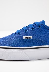 Sneaker blu glitterate con tomaia in tessuto, chiusura con lacci, suola in gomma bianca e strisce nere sui lati. Il logo Vans è ben visibile.