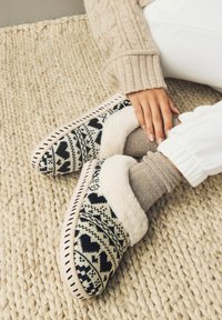 Chaussons tricotés noirs et crème avec une doublure en fourrure, présentant un motif en cœur et des surpiqûres décoratives sur un tapis texturé.