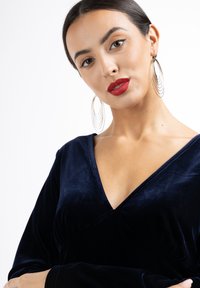 Rochie drapată din catifea albastră marină, cu decolteu adânc în V și mâneci lungi. Modelul poartă cercuri mari din argint și ruj roșu îndrăzneț. Textură fină.