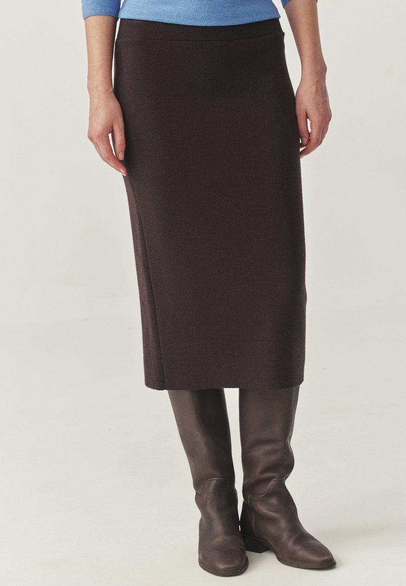 Femme portant une jupe marron foncé longueur genou, un haut bleu et de hautes bottes en cuir marron foncé, debout devant un fond clair uni.