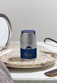 NIVEA Men Anti-Age Hyaluron cream in a silver and blue bottle with a round cap. Placed on a speckled stone tray beside a mirror and tools.