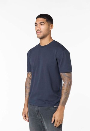 Homme tatoué portant un t-shirt bleu marine à manches courtes et un jean gris foncé, debout devant un fond blanc uni.