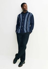 Jūrinis marškinėlių mygtukų viršus su vertikaliomis mėlynomis juostelėmis, tekstūruota medžiaga; derinamas su juodomis kelnėmis ir tamsiais batais, stovintis prieš vienodą fono.