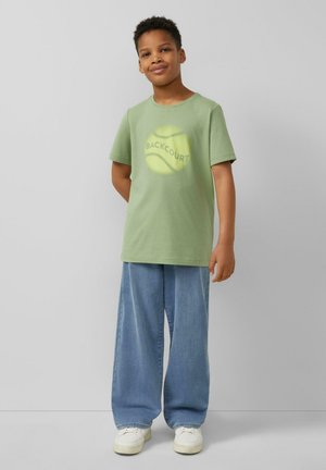 Garçon debout, les mains derrière le dos, portant un t-shirt vert « Backcourt » avec une balle de tennis, un large jean bleu et des baskets blanches, sur un fond gris.