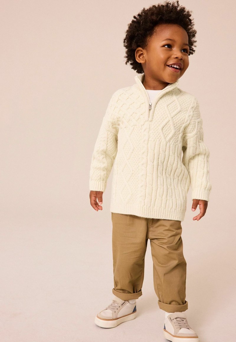 Pull en crème tricoté avec fermeture éclair, présentant des motifs texturés en losanges et en torsades. Associé à un pantalon beige clair et des baskets beiges.