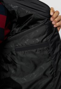 Schwarze Jackeninnenseite mit glatter Textur. Verfügt über eine versteckte Tasche, die mit dem U.S. Polo Assn. Branding in einem wiederholten Muster gefüttert ist.