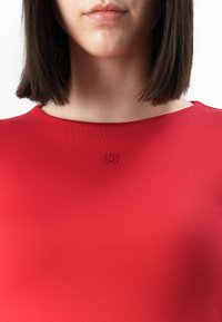 Vrouw met steil donker haar die een rode top draagt met een klein reliëf "HUGO" logo op de centrale decolletélijn, tegen een witte achtergrond.
