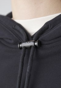 Sweat à capuche noir avec cordons ajustables retenus par un arrêt en plastique noir avec l’inscription « NAPAPIJRI » au-dessus d’un col blanc.
