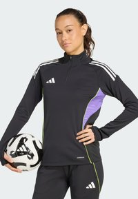 Crni sportski gornji dio s četvrtastim zipom, bijelim prugama na ramenima i ljubičastim bočnim panelima. Materijal izgleda rastezljivo. Model drži crno-bijelu nogometnu loptu.