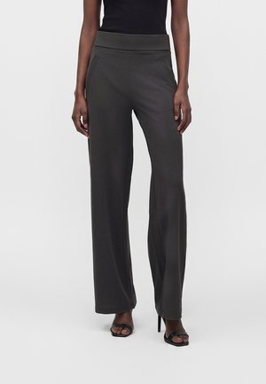 Persoon draagt een donkergrijze broek met wijde pijpen, een zwarte mouwloze top en zwarte sandalen met hak, staande tegen een effen lichte achtergrond.