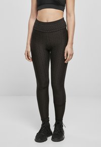 Leggings noirs taille haute avec un motif hexagonal texturé, fabriqués à partir d'un tissu extensible, associés à des chaussures de sport noires.