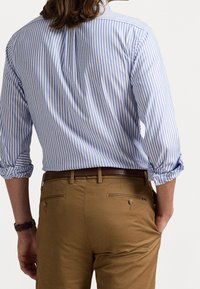 Světle modrá a bílá pruhovaná košile s ohrnutými rukávy, nošená přes hnědé chinos kalhoty s zadními kapsami a knoflíkovými detaily.