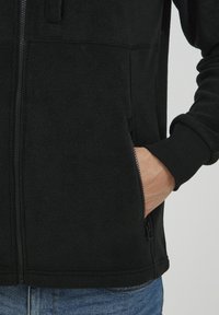 Mustassa fleece-takissa on vetoketju ja sivutaskut. Takissa on pehmeä rakenne ja ribbireunukset, jotka on suunniteltu mukavuutta ja lämpöä varten.