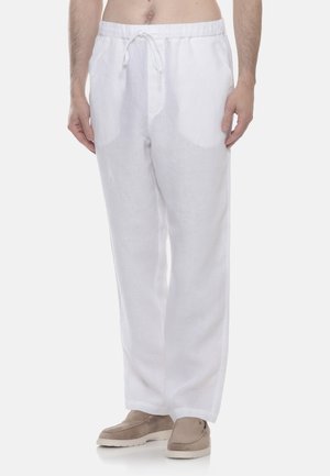 Uomo con pantaloni larghi di lino bianchi con coulisse in vita e scarpe beige slip-on, in piedi davanti a uno sfondo semplice.