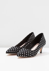 Black pointed-toe heels with white polka dots and short, beige stiletto heels, shown side by side on a white surface.
