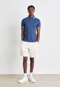 Blå polo skjorte med hvide detaljer, cremefarvede shorts og hvide sneakers. Poloen har et lille logo og en krave med knapper.
