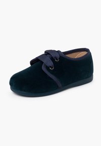 Pantofola in velluto verde scuro con punta rotonda, lacci in nastro blu navy e soletta imbottita. Suola piatta e flessibile per il massimo comfort.