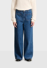 Osoba nosi široke plave traperice s velikim prednjim džepovima, crne cipele i bijeli pleteni pulover, stojeći na jednostavnoj podlozi.