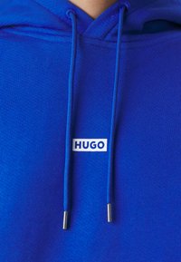 Blå hettegenser med teksturert stoff, med en hvit rektangulær logo merket "HUGO." Inkluderer to matchende blå snorer med metalltips.