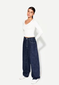 Vit långärmad V-hals topp tillsammans med vida mörkblå denimjeans. Jeansen har synlig sömnad och knappstängning, kompletterat med vita sneakers.