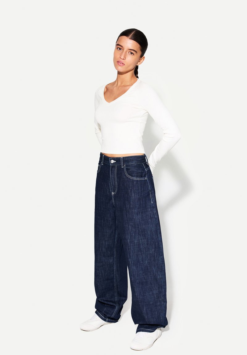 Vit långärmad V-hals topp tillsammans med vida mörkblå denimjeans. Jeansen har synlig sömnad och knappstängning, kompletterat med vita sneakers.