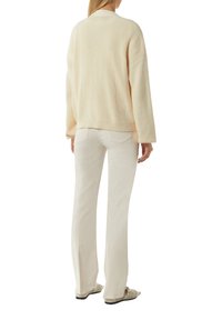 Helle beige Strickpullover mit Rippstruktur, oversized Schnitt und gesenkten Schultern. Kombiniert mit crèmefarbenen Schlaghosen und Slippern.