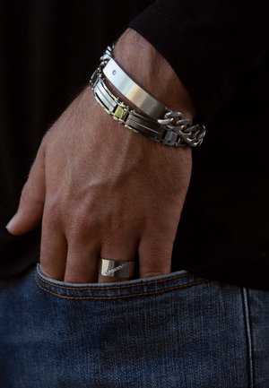Zilveren metalen armband met meerdere schakels, gecombineerd met een platte band. Vinger draagt een gepolijste ring die tegen een denim stof rust.