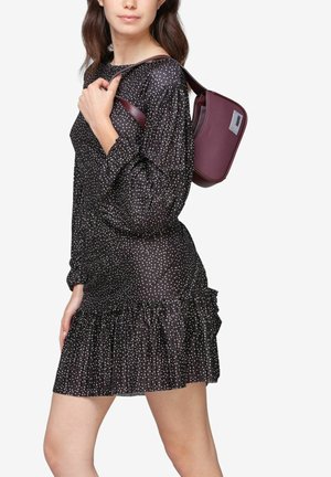 Zwarte jurk met witte stippen, lange mouwen en een gerimpelde zoom. Model draagt een bordeauxrode crossbody tas met een slank ontwerp en logo-detail.