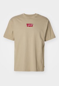 Krémové tričko s krátkým rukávem a kulatým výstřihem s červeným logem Levi’s umístěným na středu hrudi na jednobarevném pozadí.