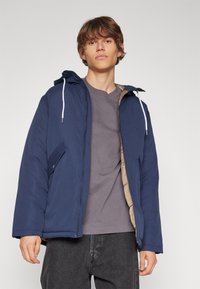 Doudoune bleu marine avec capuche, dotée d'une doublure beige et de cordons blancs. Comprend une poche latérale et un design à coupe décontractée.