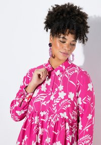Rosa Blüten-Hemdblusenkleid mit weißen Blumenmustern, gerafftem Taillenbund und langen Ärmeln mit blumigen Manschetten. Große runde Ohrringe schmücken das Model.