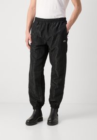Schwarze, leichte Jogginghosen mit einem elastischen Bund, schmal zulaufenden Säumen und einer dezenten Textur, die ein kleines grünes Logo auf der linken Seite tragen.
