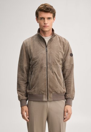 Jonge man met kort bruin haar die een taupekleurige suède jas met rits en beige broek draagt tegen een effen lichte achtergrond.