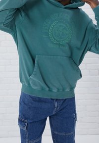 Felpa con cappuccio color teal e tasca frontale, decorata con grafiche verdi stampate. Abbinata a pantaloni cargo in denim blu con molteplici tasche.