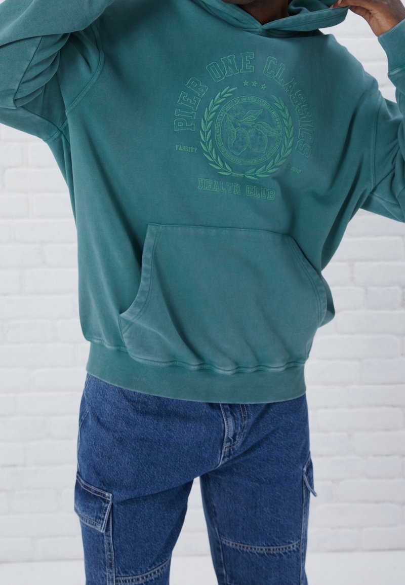 Felpa con cappuccio color teal e tasca frontale, decorata con grafiche verdi stampate. Abbinata a pantaloni cargo in denim blu con molteplici tasche.
