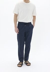 Pantalones elásticos de talle medio, ligeros y de color azul marino, con bolsillos laterales, combinados con una camiseta blanca de manga corta y sandalias oscuras.