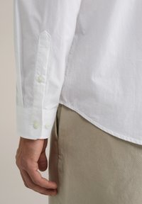 Witte blouse met lange mouwen en een spitse manchette, met drie lichte knopen. De stof lijkt glad en lichtjes gestructureerd. Tan broek zichtbaar.