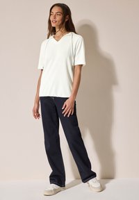 Hodie blanc, texturé, à manches courtes, avec un col en V et des cordons de serrage, associé à un jean bleu foncé et des baskets blanches. Coupe simple et décontractée.