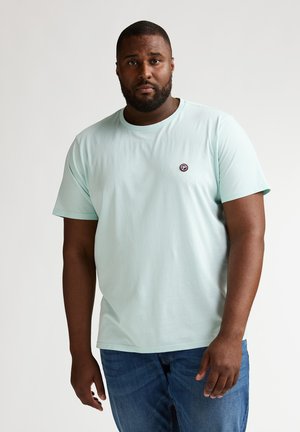 Lichtblauw katoenen T-shirt met korte mouwen, ronde hals en een klein rond logoplaatje op de linkerborst; gecombineerd met een blauwe spijkerbroek.