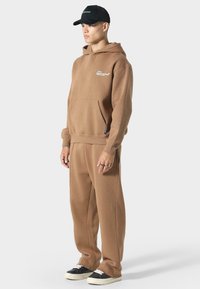 Brun hættetrøje og matchende sweatpants lavet af blødt stof. Hættetrøjen har en frontlomme og et diskret logo. Accessories inkluderer en sort kasket.