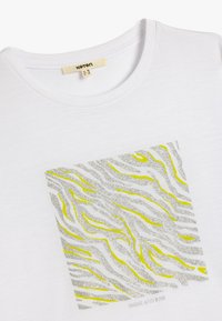 Camiseta blanca de algodón con un gráfico cuadrado que presenta rayas de cebra en plata y amarillo. El texto dice "BRILLA Y SURGE" en la parte inferior.