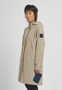 Beige long coat with a hood, front zipper, and elastic cuffs. Features a black patch on the sleeve and smooth, lightweight fabric.