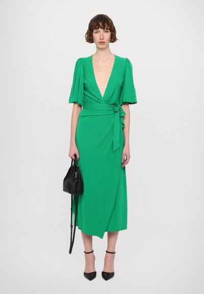 Green wrap dress with short puff sleeves, deep V-neck, and tie at waist. Model holds a black bag, wearing pointed black heels.